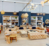 A stylish three-seat brown leather Chesterfield sofa with a modern design, featuring deep button tufting and rolled arms, placed in a bright, contemporary living space. The background includes shelves filled with decorative items and plants, along with a warm color palette and ambient lighting