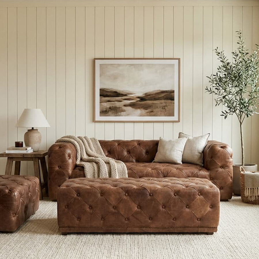 Brown leather rectangle Chesterfield ottoman placed in a cozy living room setting, accompanied by a matching brown leather sofa and a small side table with a lamp. A large piece of wall art hangs above the sofa, and a potted plant adds a touch of greenery to the space. The floor is covered with a light-colored rug, complementing the warm tones of the furniture and wooden flooring