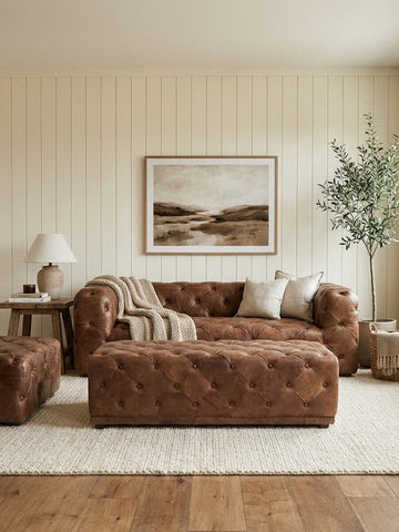 Brown leather rectangle Chesterfield ottoman placed in a cozy living room setting, accompanied by a matching brown leather sofa and a small side table with a lamp. A large piece of wall art hangs above the sofa, and a potted plant adds a touch of greenery to the space. The floor is covered with a light-colored rug, complementing the warm tones of the furniture and wooden flooring