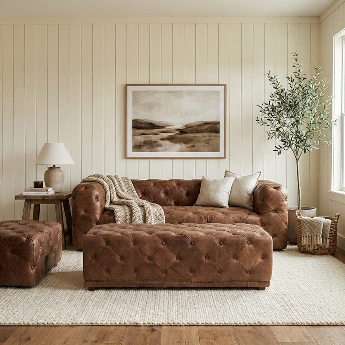 A brown leather rectangle Chesterfield ottoman sits in a cozy living room, accompanied by a matching brown leather sofa. The room features light-colored walls, a large framed landscape artwork, a potted plant, and a lamp on a side table, all arranged on a soft area rug. Natural light floods in from a window, creating a warm and inviting atmosphere