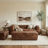 A brown leather rectangle Chesterfield ottoman sits in a cozy living room, accompanied by a matching brown leather sofa. The room features light-colored walls, a large framed landscape artwork, a potted plant, and a lamp on a side table, all arranged on a soft area rug. Natural light floods in from a window, creating a warm and inviting atmosphere