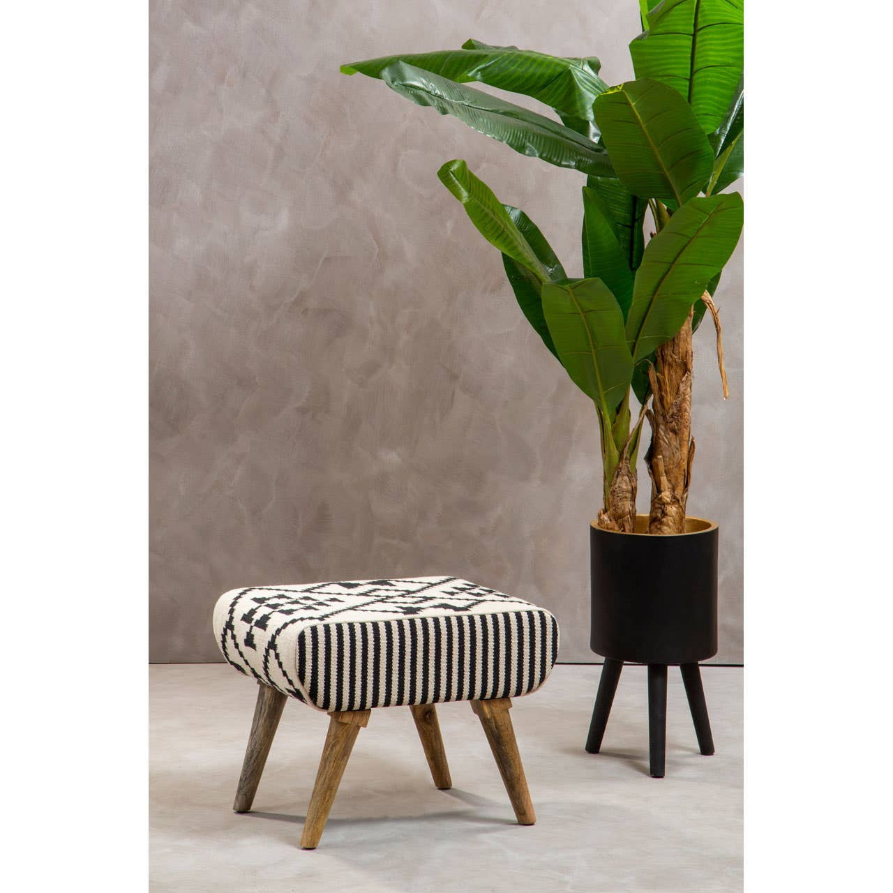 A textured footstool with a patterned fabric top in neutral tones, supported by wooden legs. It is positioned next to a tall potted plant with large green leaves against a light gray wall