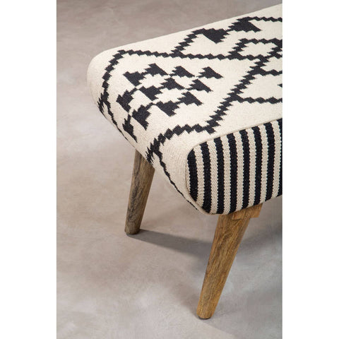 A close-up view of the Cefena Berber Style Footstool featuring a patterned fabric in cream with black geometric designs and stripes. The footstool has wooden legs that provide a natural finish, complementing the textile design. The background is a soft, neutral surface