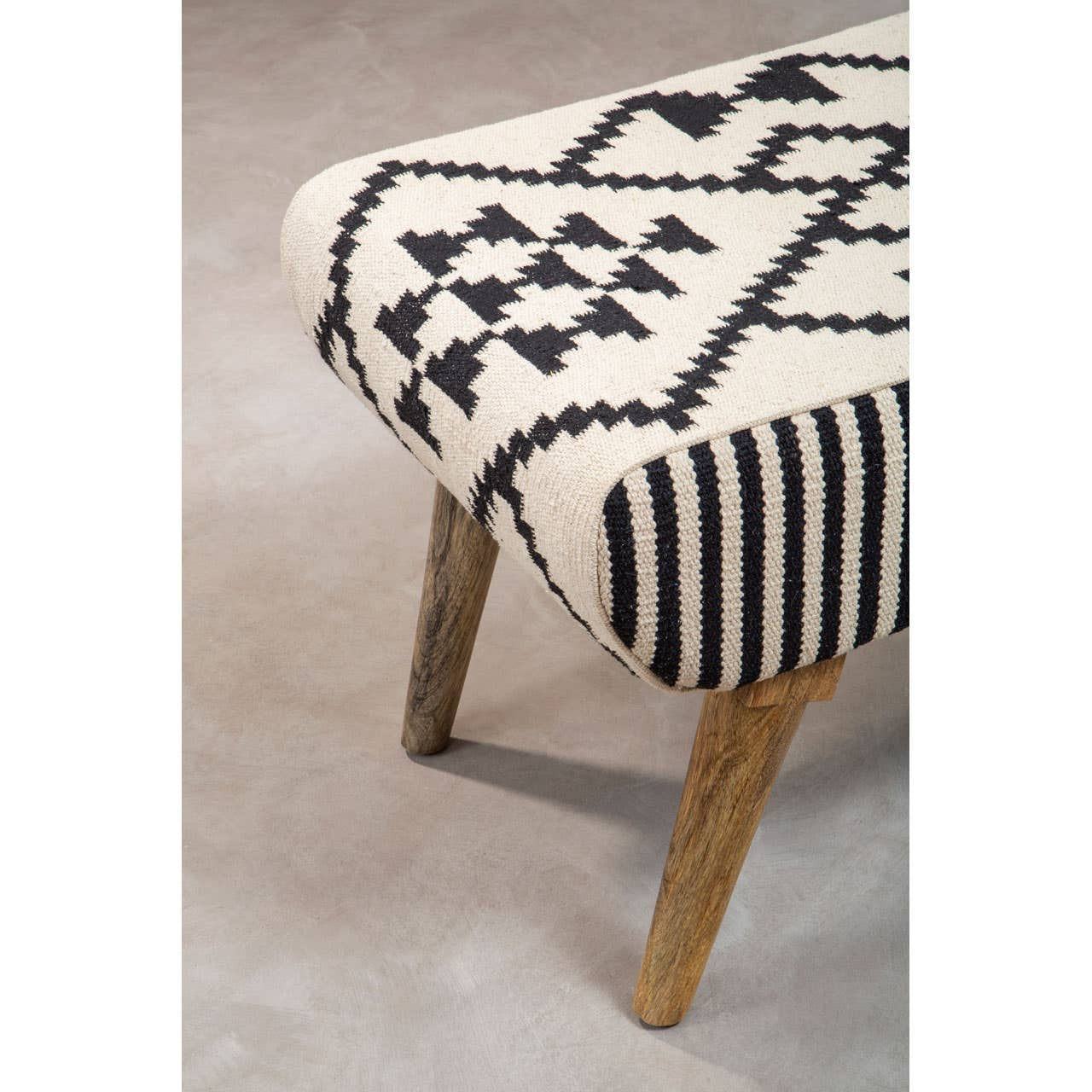 A close-up view of the Cefena Berber Style Footstool featuring a patterned fabric in cream with black geometric designs and stripes. The footstool has wooden legs that provide a natural finish, complementing the textile design. The background is a soft, neutral surface