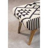 A close-up view of the Cefena Berber Style Footstool featuring a patterned fabric in cream with black geometric designs and stripes. The footstool has wooden legs that provide a natural finish, complementing the textile design. The background is a soft, neutral surface