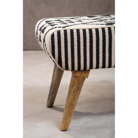 Cefena Berber Style Footstool featuring a striped fabric upholstery in black and cream, with rounded edges and wooden legs that provide a natural finish. The footstool is positioned on a light-colored floor, highlighting its design and texture