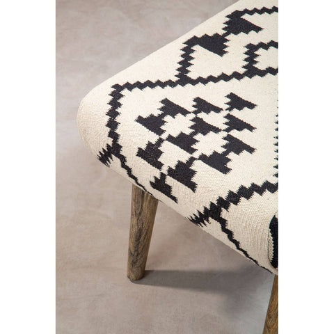 A close-up view of a Cefena Berber Style Footstool featuring a patterned fabric in cream and black with geometric designs, supported by wooden legs. The footstool is placed on a light-colored floor, highlighting its texture and design
