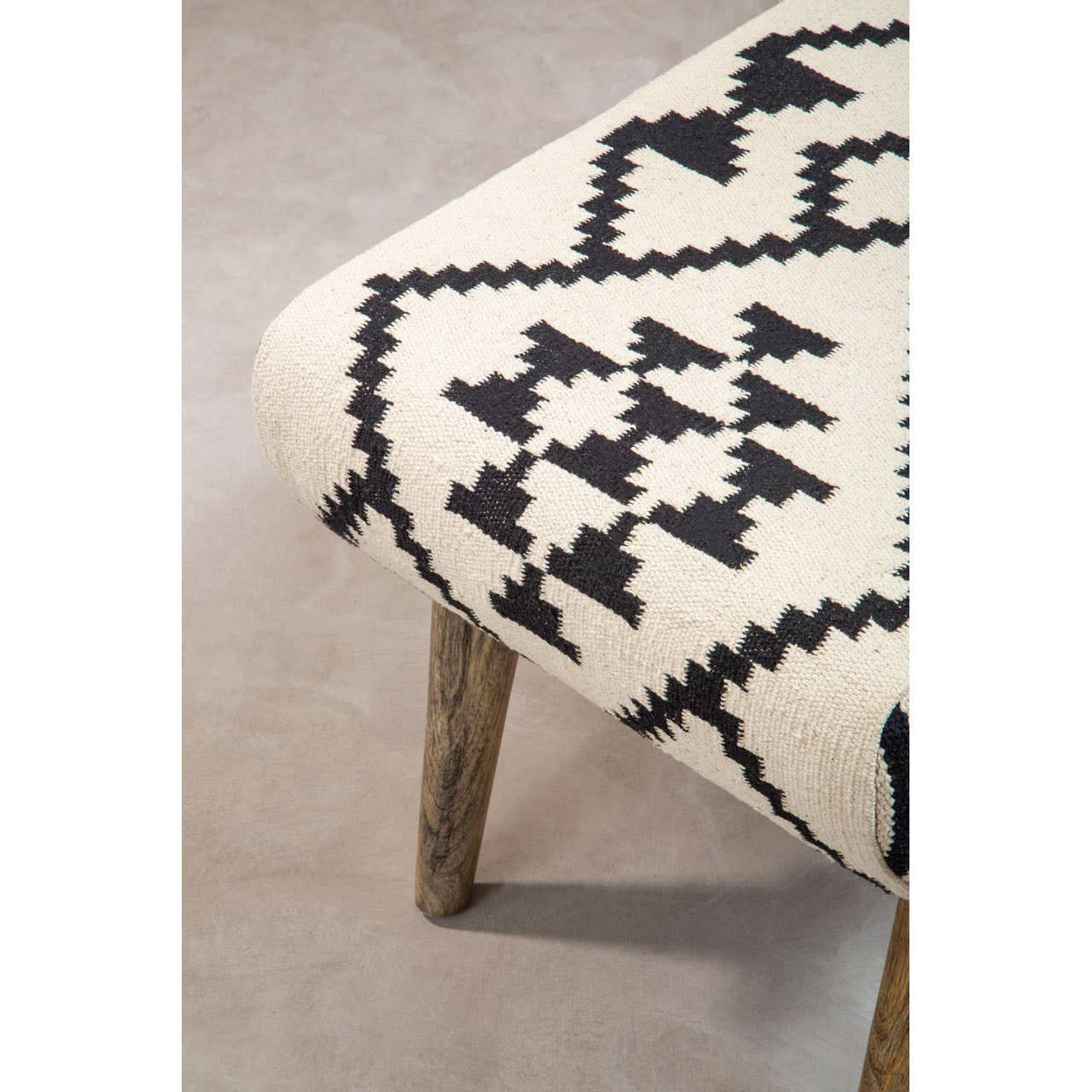 A close-up view of a Cefena Berber Style Footstool featuring a patterned fabric in cream and black with geometric designs, supported by wooden legs. The footstool is placed on a light-colored floor, highlighting its texture and design