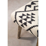 A close-up view of a Cefena Berber Style Footstool featuring a patterned fabric in cream and black with geometric designs, supported by wooden legs. The footstool is placed on a light-colored floor, highlighting its texture and design