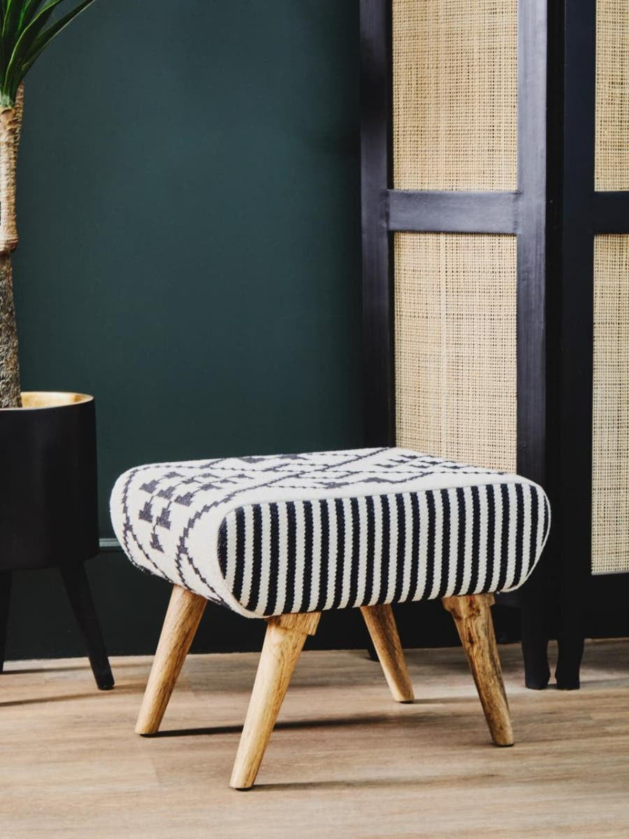 A stylish footstool with a textured gray and white fabric upholstery featuring a ribbed design, supported by four light wooden legs. The footstool is placed on a neutral-toned floor, with a dark green wall and a woven screen in the background, creating a cozy and modern atmosphere