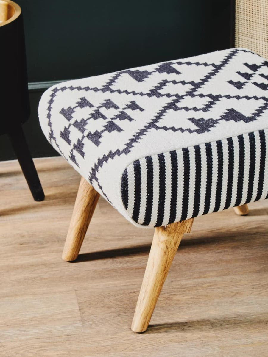 A stylish footstool featuring a patterned fabric in black and white with geometric designs, supported by light wooden legs. The footstool is placed on a wooden floor, showcasing its modern and cozy aesthetic