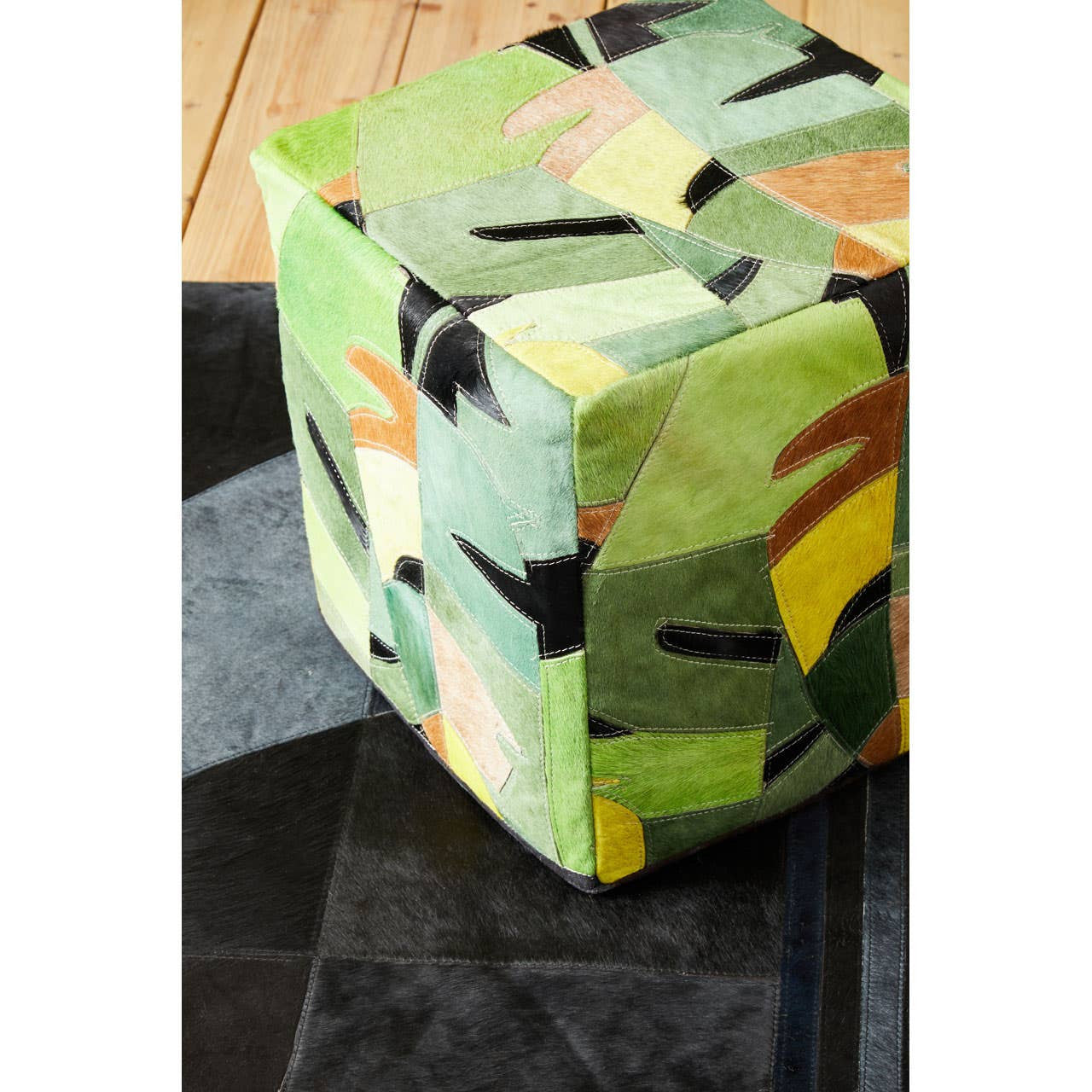 A colorful pouffe featuring a botanical design with various shades of green, yellow, and orange, set against a textured black surface. The pouffe has a cube shape and is made of leather, showcasing a vibrant, nature-inspired pattern