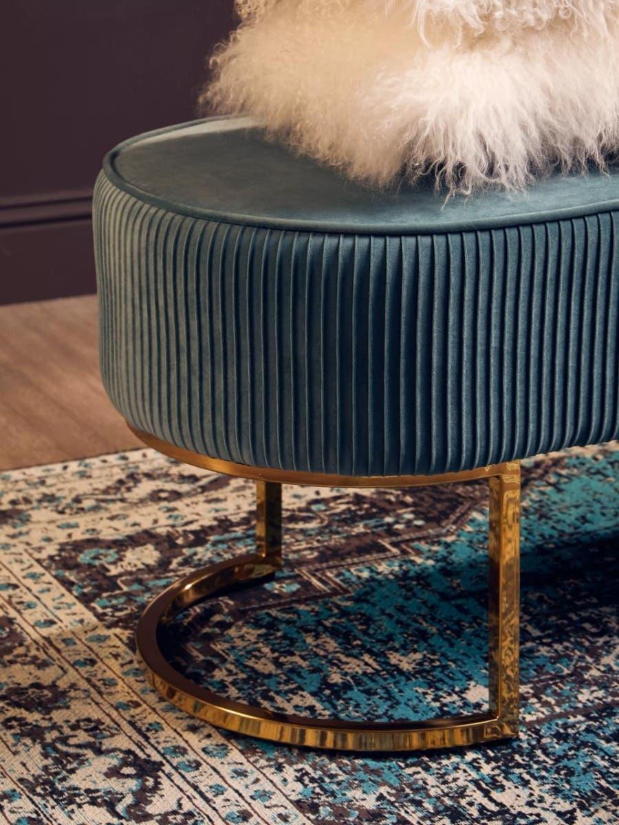 Teal velvet ottoman with gold metal base on patterned rug, topped with white faux fur cushion