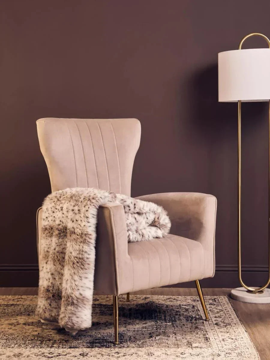 Beige accent chair with gold legs and faux fur throw on patterned rug beside floor lamp