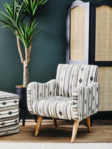 A square pouffe with a patterned fabric design sits next to a stylish armchair, both featuring light wooden legs. The background includes a dark green wall and a decorative screen with a woven texture, alongside a potted plant adding a touch of greenery to the scene
