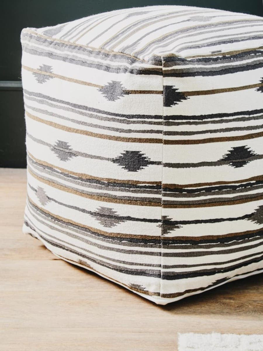 Square pouffe featuring a textured fabric with alternating stripes in cream and dark gray, showcasing a geometric pattern. The pouffe is placed on a light wooden surface, with a dark green wall in the background