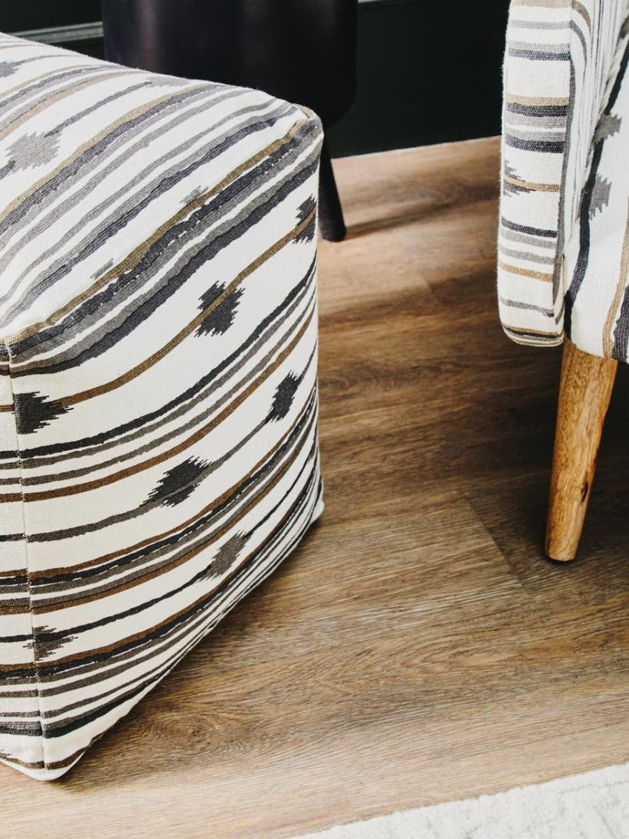 Square pouffe with a textured fabric featuring a wavy striped pattern in neutral tones, resting on a wooden floor. The pouffe has rounded edges and is supported by light wooden legs. A portion of a patterned chair is visible in the background