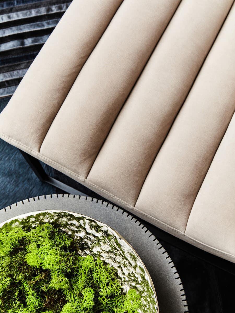 Beige upholstered bench seat with channel stitching beside decorative green moss bowl on table