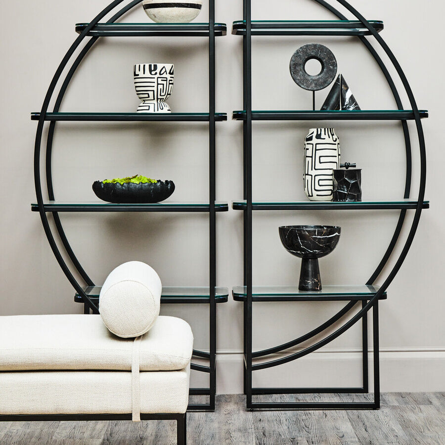 A black half-moon shelf unit with a circular design, featuring multiple open shelves displaying decorative items such as a white vase, a black bowl, and a small sculpture. The shelf unit is positioned against a light-colored wall, with a round white cushion resting on a nearby sofa. The floor is wooden, adding warmth to the modern aesthetic