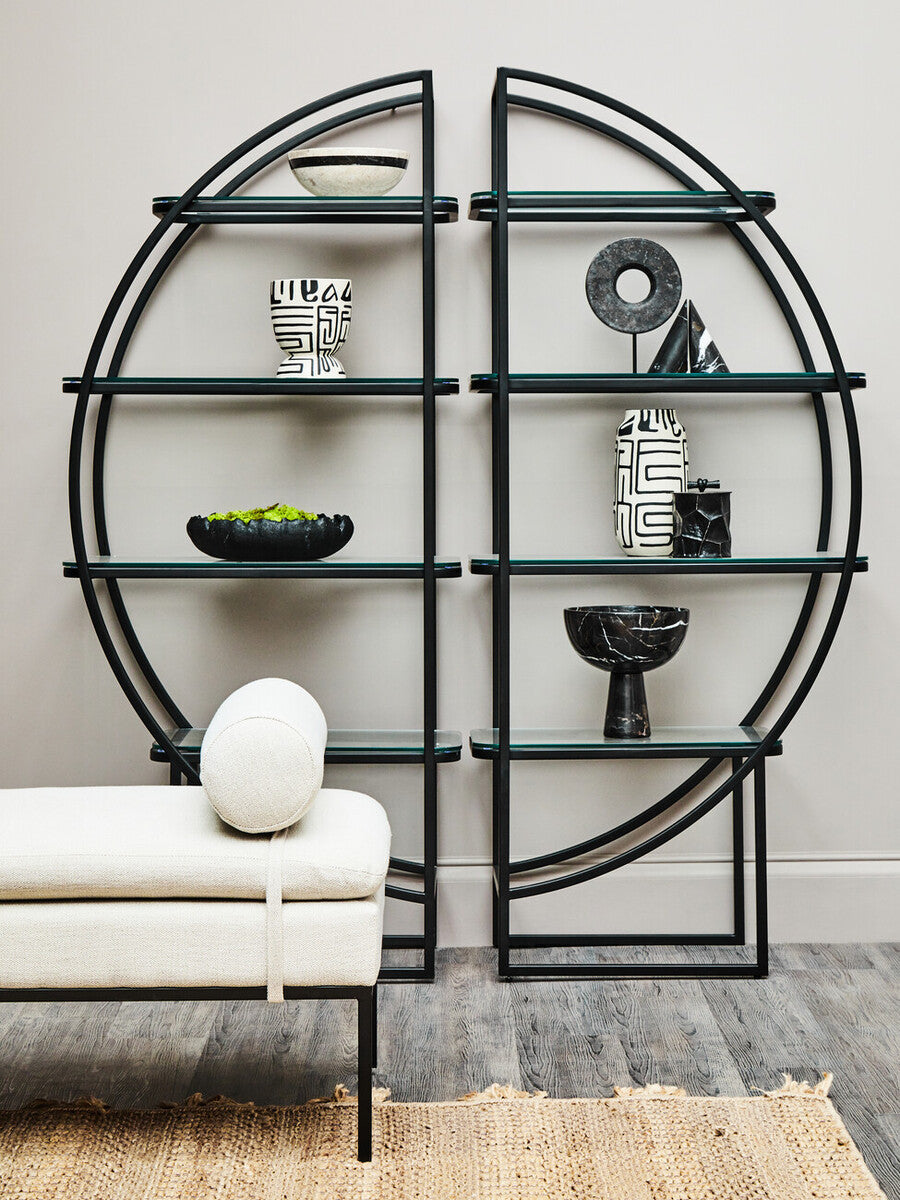 A black half-moon shelf unit with a circular design, featuring multiple open shelves displaying decorative items such as a white vase, a black bowl, and a small sculpture. The shelf unit is positioned against a light-colored wall, with a round white cushion resting on a nearby sofa. The floor is wooden, adding warmth to the modern aesthetic