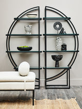 A black half-moon shelf unit with a circular design, featuring multiple open shelves displaying decorative items such as a white vase, a black bowl, and a small sculpture. The shelf unit is positioned against a light-colored wall, with a round white cushion resting on a nearby sofa. The floor is wooden, adding warmth to the modern aesthetic