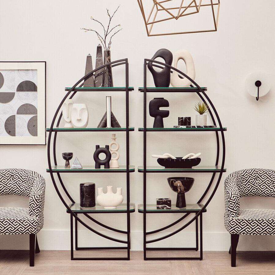 A black half-moon shaped shelf unit with a geometric design, featuring multiple open shelves displaying decorative items such as vases, books, and sculptures. Two light gray upholstered chairs are positioned in front of the shelf unit, and a modern light fixture hangs above. The background includes framed artwork on the wall