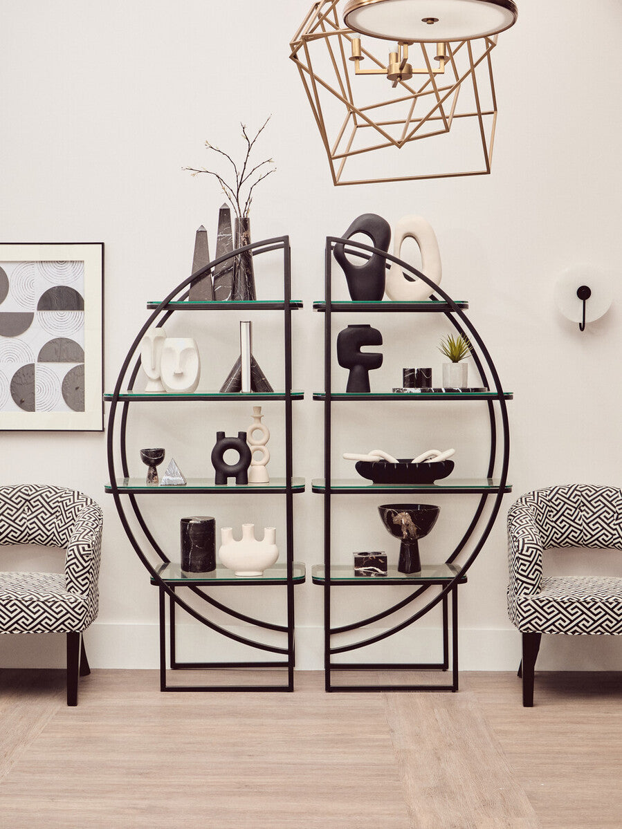 A black half-moon shaped shelf unit with a geometric design, featuring multiple open shelves displaying decorative items such as vases, books, and sculptures. Two light gray upholstered chairs are positioned in front of the shelf unit, and a modern light fixture hangs above. The background includes framed artwork on the wall