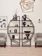 A black half-moon shaped shelf unit with a geometric design, featuring multiple open shelves displaying decorative items such as vases, books, and sculptures. Two light gray upholstered chairs are positioned in front of the shelf unit, and a modern light fixture hangs above. The background includes framed artwork on the wall