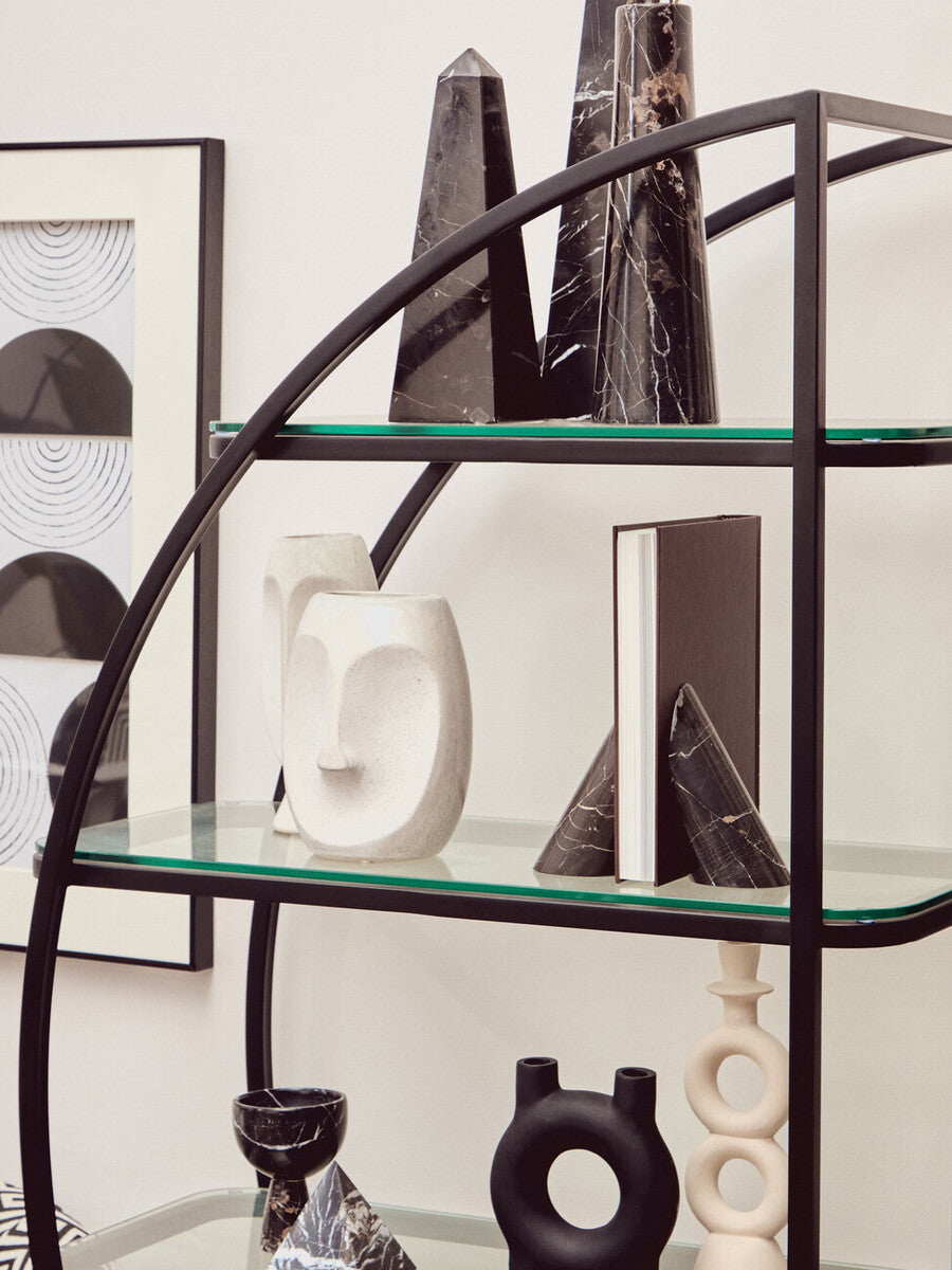 A black half-moon shelf unit featuring a sleek metal frame with multiple glass shelves. The shelves display various decorative items, including abstract sculptures and vases, against a neutral wall backdrop