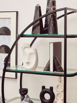 A black half-moon shelf unit featuring a sleek metal frame with multiple glass shelves. The shelves display various decorative items, including abstract sculptures and vases, against a neutral wall backdrop
