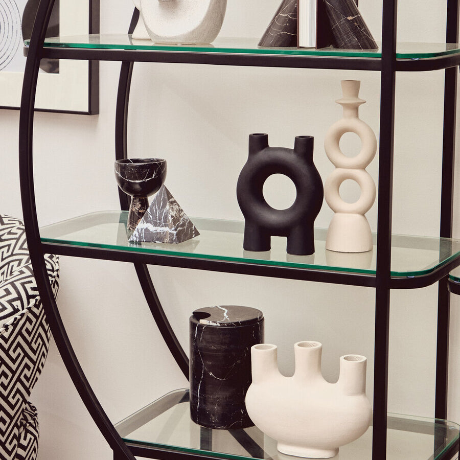 A black half-moon shelf unit with a sleek metal frame and multiple glass shelves, displaying various decorative items including black and white vases and sculptures. The background features a light-colored wall with framed artwork