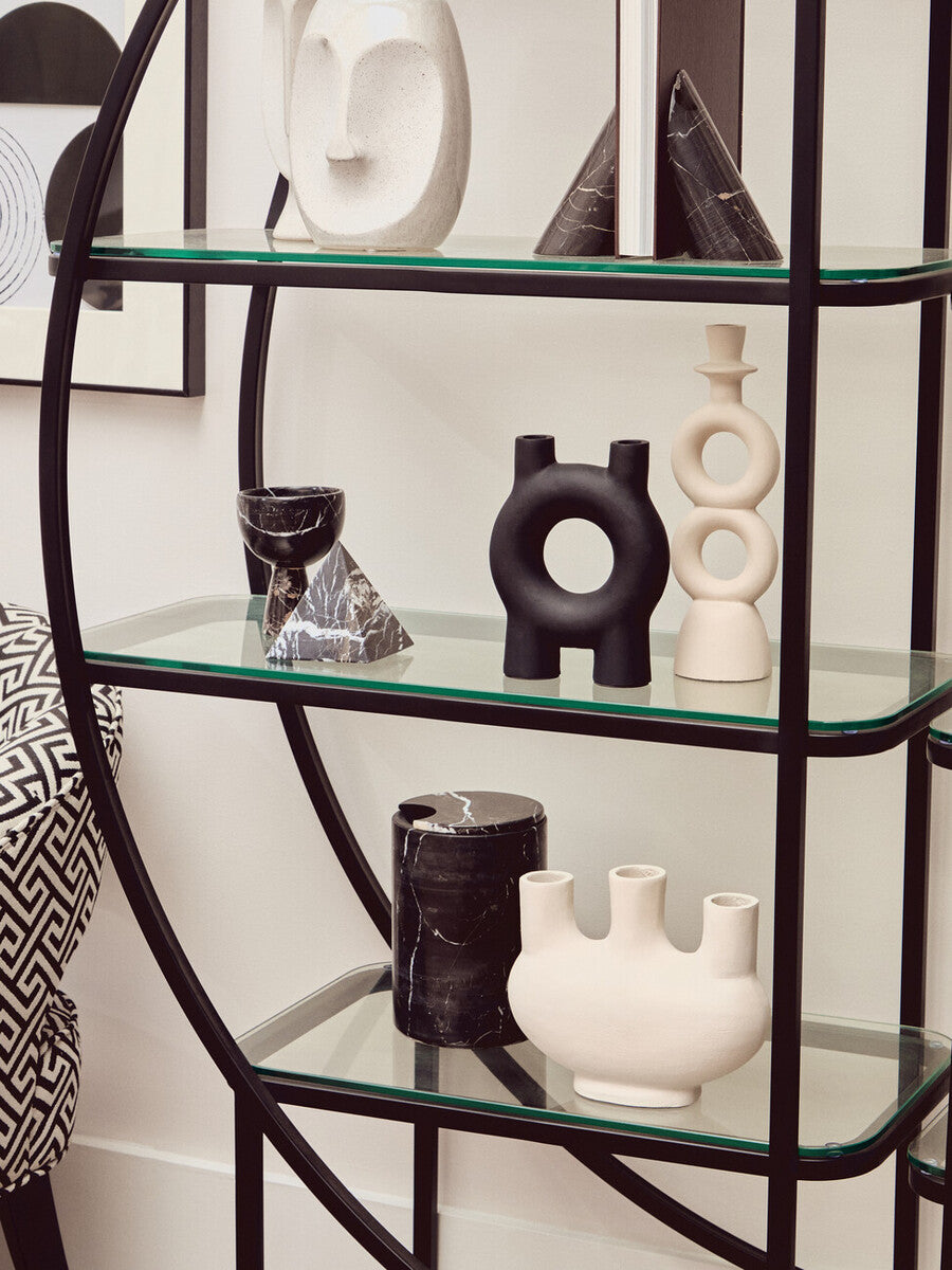 A black half-moon shelf unit with a sleek metal frame and multiple glass shelves, displaying various decorative items including black and white vases and sculptures. The background features a light-colored wall with framed artwork
