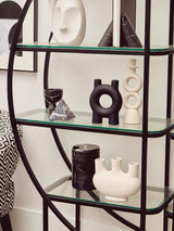 A black half-moon shelf unit with a sleek metal frame and multiple glass shelves, displaying various decorative items including black and white vases and sculptures. The background features a light-colored wall with framed artwork