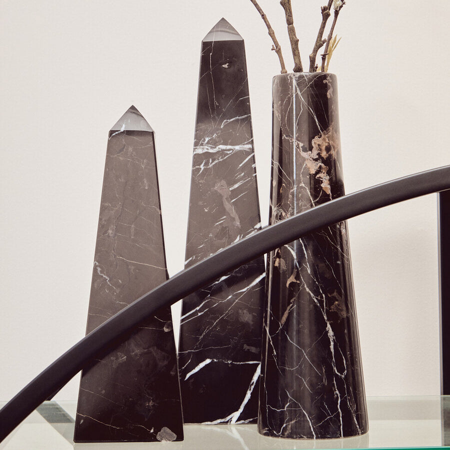 A black shelf unit with a half-moon shape, featuring three tall, black decorative obelisks and a slender vase with branches, set against a light-colored wall. The shelf has a sleek design with a glass top and a curved black frame