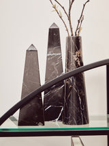 A black shelf unit with a half-moon shape, featuring three tall, black decorative obelisks and a slender vase with branches, set against a light-colored wall. The shelf has a sleek design with a glass top and a curved black frame