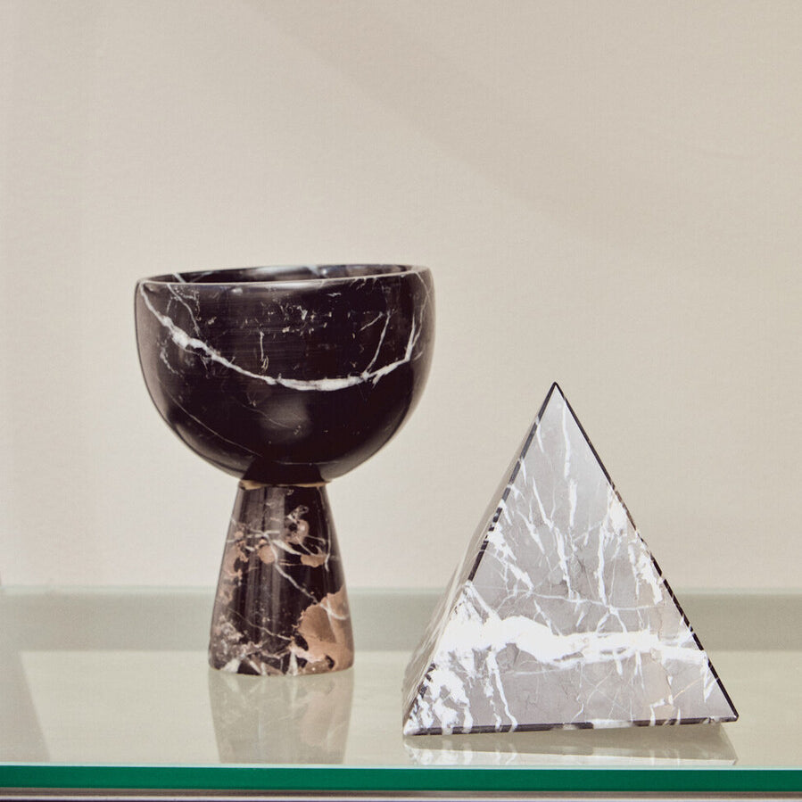 A black half-moon shelf unit displaying a textured black vase with a curved top and a decorative silver pyramid sculpture, set against a neutral background