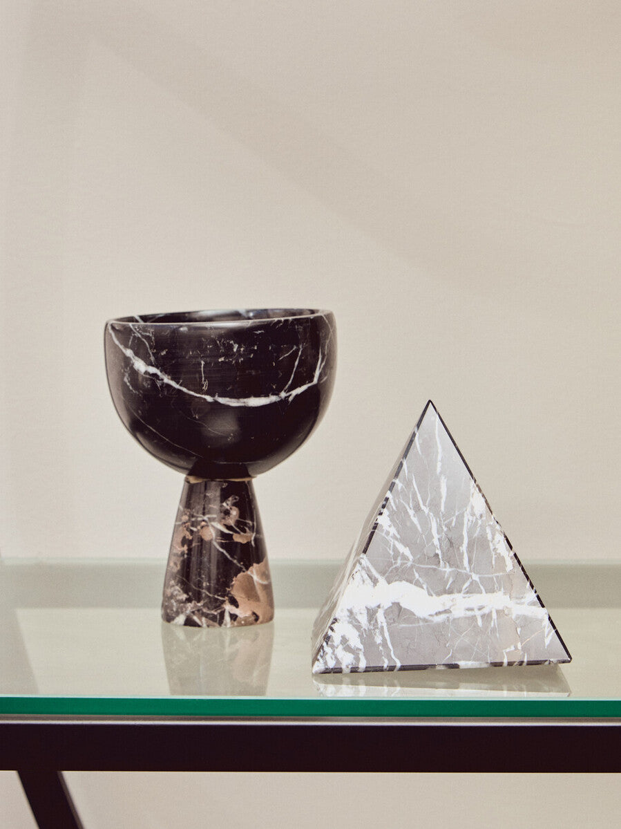 A black half-moon shelf unit displaying a textured black vase with a curved top and a decorative silver pyramid sculpture, set against a neutral background