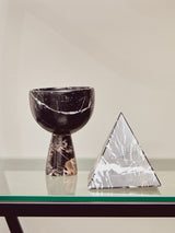A black half-moon shelf unit displaying a textured black vase with a curved top and a decorative silver pyramid sculpture, set against a neutral background