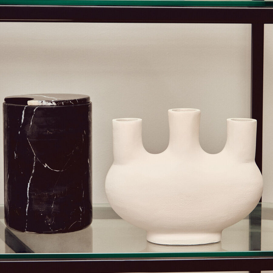 A black shelf unit with a left half-moon design, featuring a black cylindrical vase and a white ceramic vase with three openings, both placed on the shelves. The background is a light-colored wall, enhancing the modern aesthetic of the display