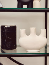 A black shelf unit with a left half-moon design, featuring a black cylindrical vase and a white ceramic vase with three openings, both placed on the shelves. The background is a light-colored wall, enhancing the modern aesthetic of the display
