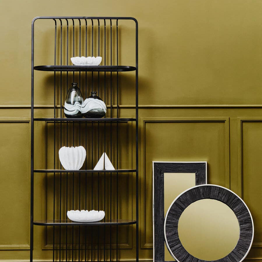 A five-tier black shelf unit with an open design, featuring decorative items such as white vases and a geometric sculpture on the shelves. The background is a warm yellow wall, and there are two round mirrors leaning against the wall, one with a black frame and the other with a gold finish. A patterned rug is visible on the floor