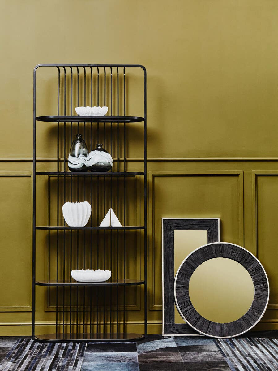 A five-tier black shelf unit with an open design, featuring decorative items such as white vases and a geometric sculpture on the shelves. The background is a warm yellow wall, and there are two round mirrors leaning against the wall, one with a black frame and the other with a gold finish. A patterned rug is visible on the floor
