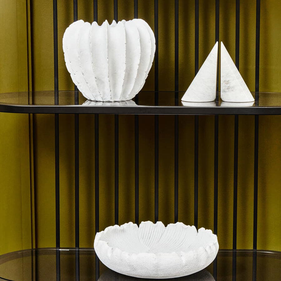 A five-tier black shelf unit featuring sleek, modern lines. The shelves display three white decorative items: a textured bowl, a fluted vase, and a triangular sculpture, all set against a warm yellow background