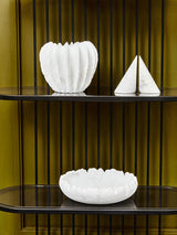 A five-tier black shelf unit featuring sleek, modern lines. The shelves display three white decorative items: a textured bowl, a fluted vase, and a triangular sculpture, all set against a warm yellow background