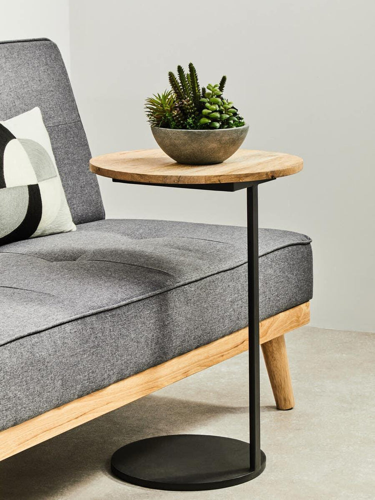 Round wooden side table with black metal base beside grey sofa, decorative plant on top