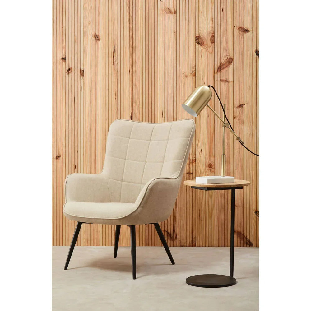 Beige upholstered armchair with black legs beside round side table and gold desk lamp