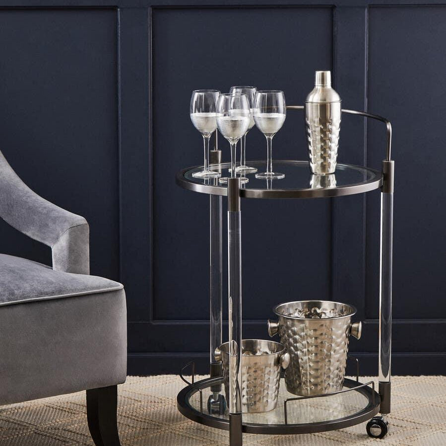Glass and metal drinks trolley with wine glasses, cocktail shaker, and ice buckets beside velvet chair
