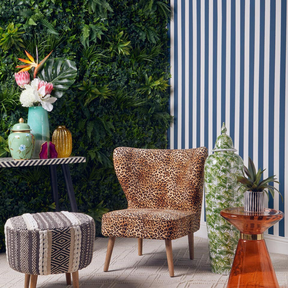 A grey and white patterned stool with a round shape sits on a light-colored floor, surrounded by a cozy chair, a small table with colorful vases, and a vibrant orange side table. Lush green plants and a striped wall provide a stylish backdrop