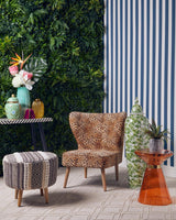 A grey and white patterned stool with a round shape sits on a light-colored floor, surrounded by a cozy chair, a small table with colorful vases, and a vibrant orange side table. Lush green plants and a striped wall provide a stylish backdrop