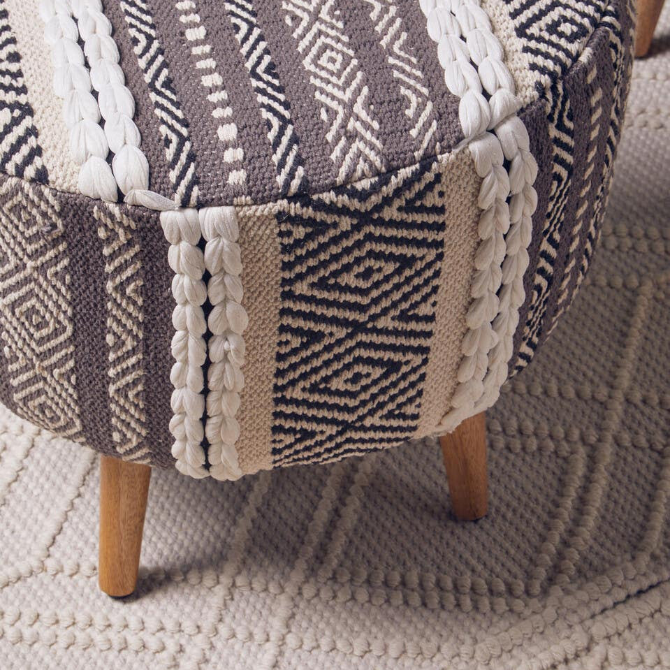 A grey and white patterned stool with a rounded top and wooden legs, placed on a textured light-colored rug. The fabric features various geometric designs in shades of grey, cream, and white